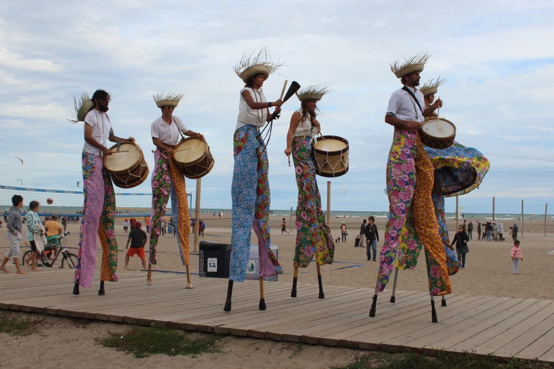 Five TallBeat stilt drummers performing Maracatu rhythms on Toronto beach boardwalk with ocean and crowd in background
